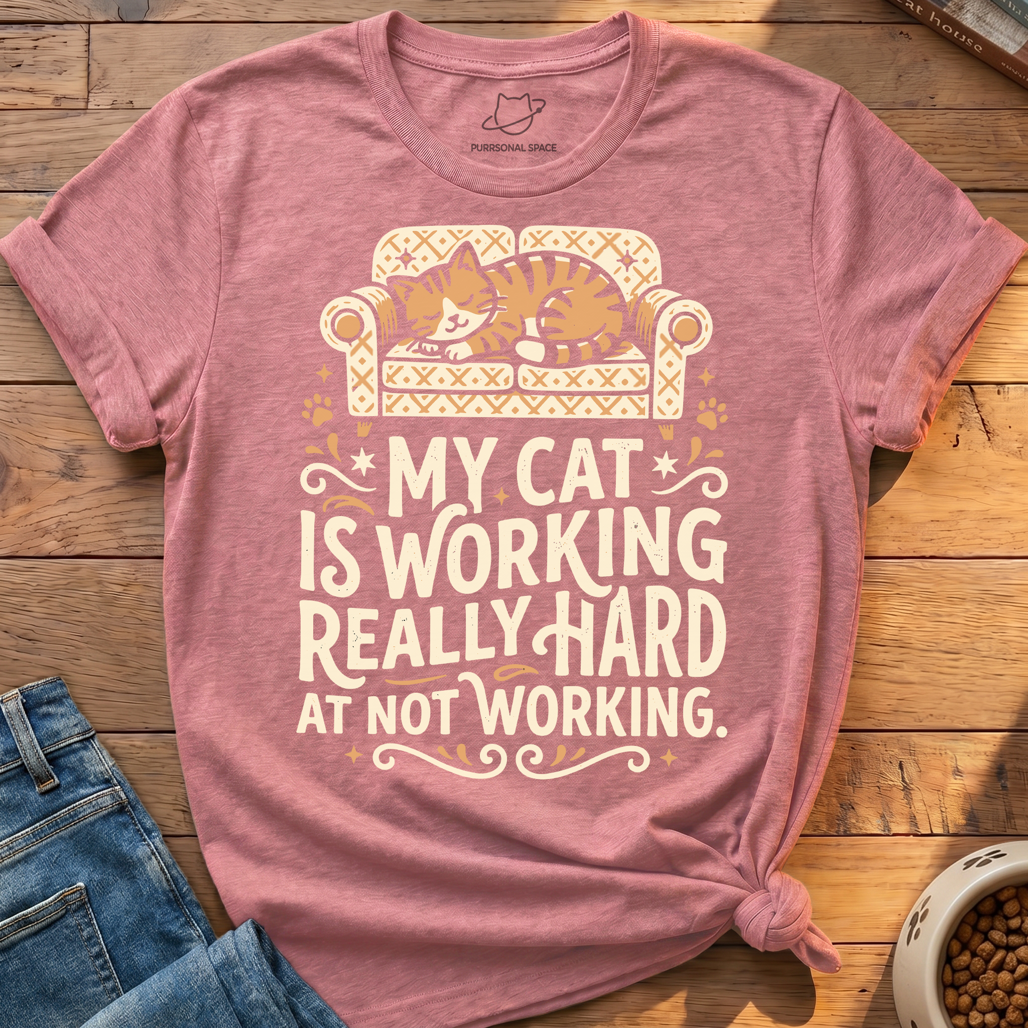 A heathered dusty rose t-shirt featuring a cute graphic of an orange tabby cat sleeping on a patterned cream sofa, with the text 'MY CAT IS WORKING REALLY HARD AT NOT WORKING.' printed below.