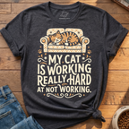 A dark heather gray graphic t-shirt featuring an illustration of a sleeping orange tabby cat on a patterned sofa with the text 'MY CAT IS WORKING REALLY HARD AT NOT WORKING.' The shirt is styled in a flat lay on a wooden floor with blue jeans and a bowl of cat food visible at the edges.