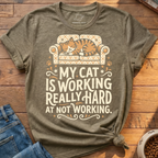 A heather olive green graphic t-shirt featuring a cartoon cat sleeping on a patterned sofa and the text 'MY CAT IS WORKING REALLY HARD AT NOT WORKING.' The shirt is styled in a flat lay on a wooden floor with blue jeans and a bowl of cat food nearby.