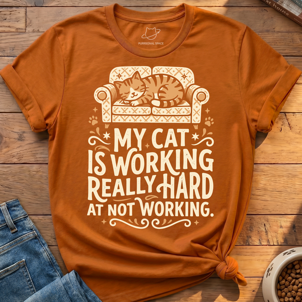 An orange short-sleeve graphic t-shirt with a cream-colored print of a cat sleeping on a patterned couch and the text 'MY CAT IS WORKING REALLY HARD AT NOT WORKING.' The shirt is styled in a flat-lay on a wooden surface next to blue jeans and a bowl of cat food.