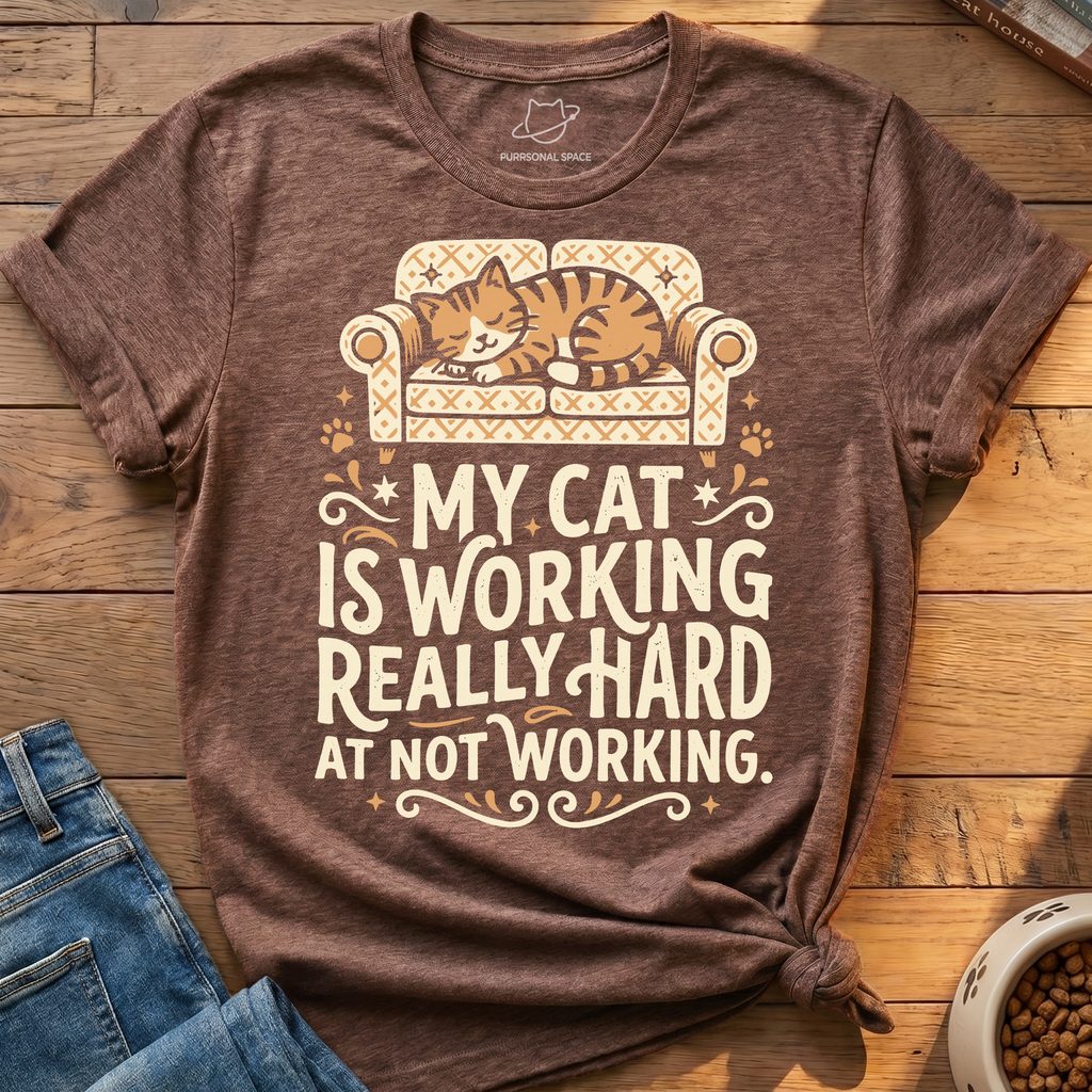 A heather brown graphic t-shirt featuring a cartoon cat sleeping on a patterned armchair with the text 'MY CAT IS WORKING REALLY HARD AT NOT WORKING.' The shirt is styled in a flat-lay on a wooden surface next to blue jeans and a bowl of cat food.
