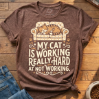 A heather brown graphic t-shirt featuring a cartoon cat sleeping on a patterned armchair with the text 'MY CAT IS WORKING REALLY HARD AT NOT WORKING.' The shirt is styled in a flat-lay on a wooden surface next to blue jeans and a bowl of cat food.