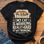 A black graphic t-shirt featuring a cartoon cat sleeping on a patterned sofa with the text 'MY CAT IS WORKING REALLY HARD AT NOT WORKING.' The shirt is styled flat on a wooden surface with blue jeans and a bowl of cat food partially visible.