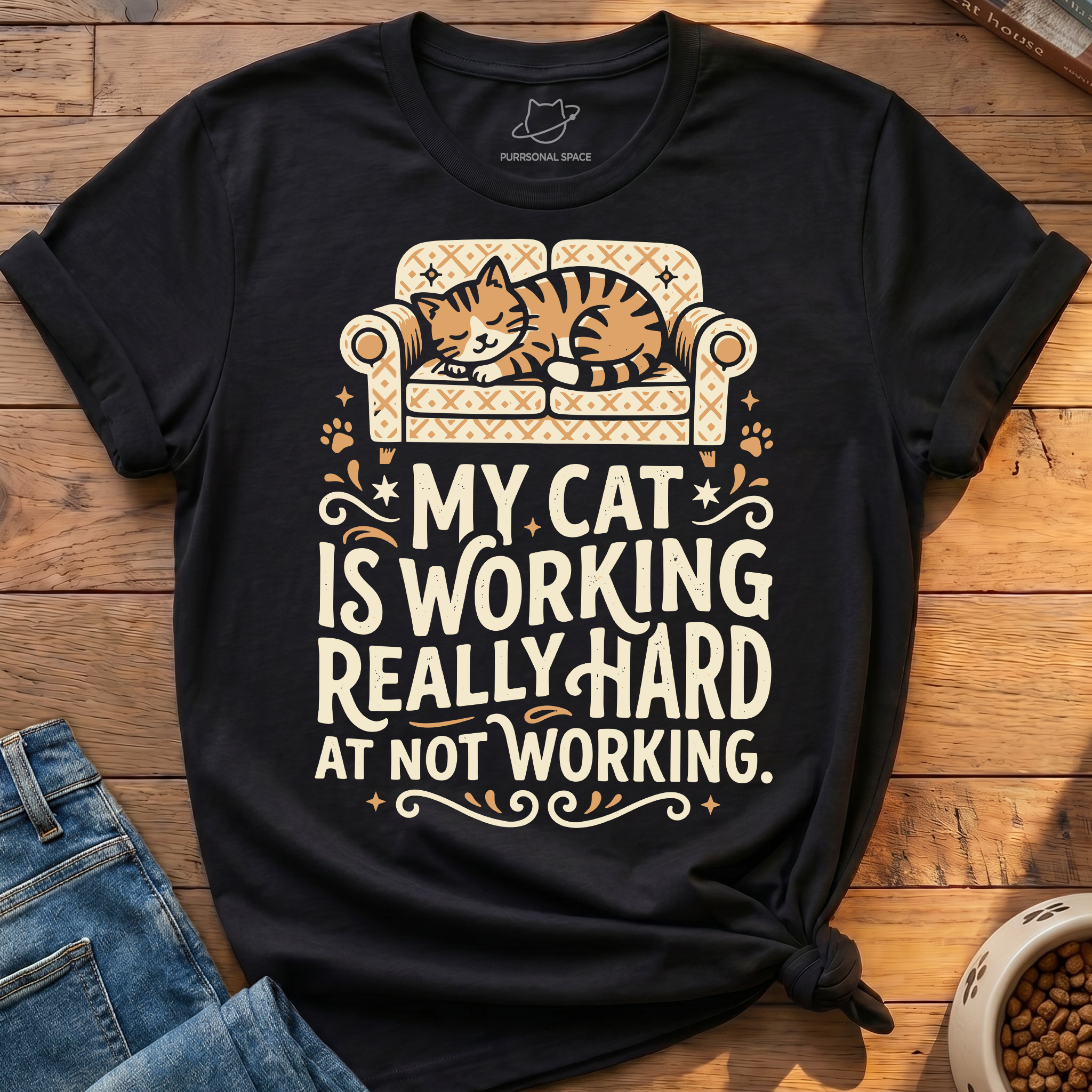 A black graphic t-shirt featuring a cartoon cat sleeping on a patterned sofa with the text 'MY CAT IS WORKING REALLY HARD AT NOT WORKING.' The shirt is styled flat on a wooden surface with blue jeans and a bowl of cat food partially visible.