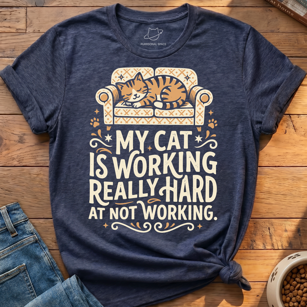 A heather navy blue graphic t-shirt featuring an illustration of a sleeping orange tabby cat on a patterned sofa with the text 'MY CAT IS WORKING REALLY HARD AT NOT WORKING.' The shirt is styled in a flat lay on a wooden floor with blue jeans and a bowl of cat food partially visible.