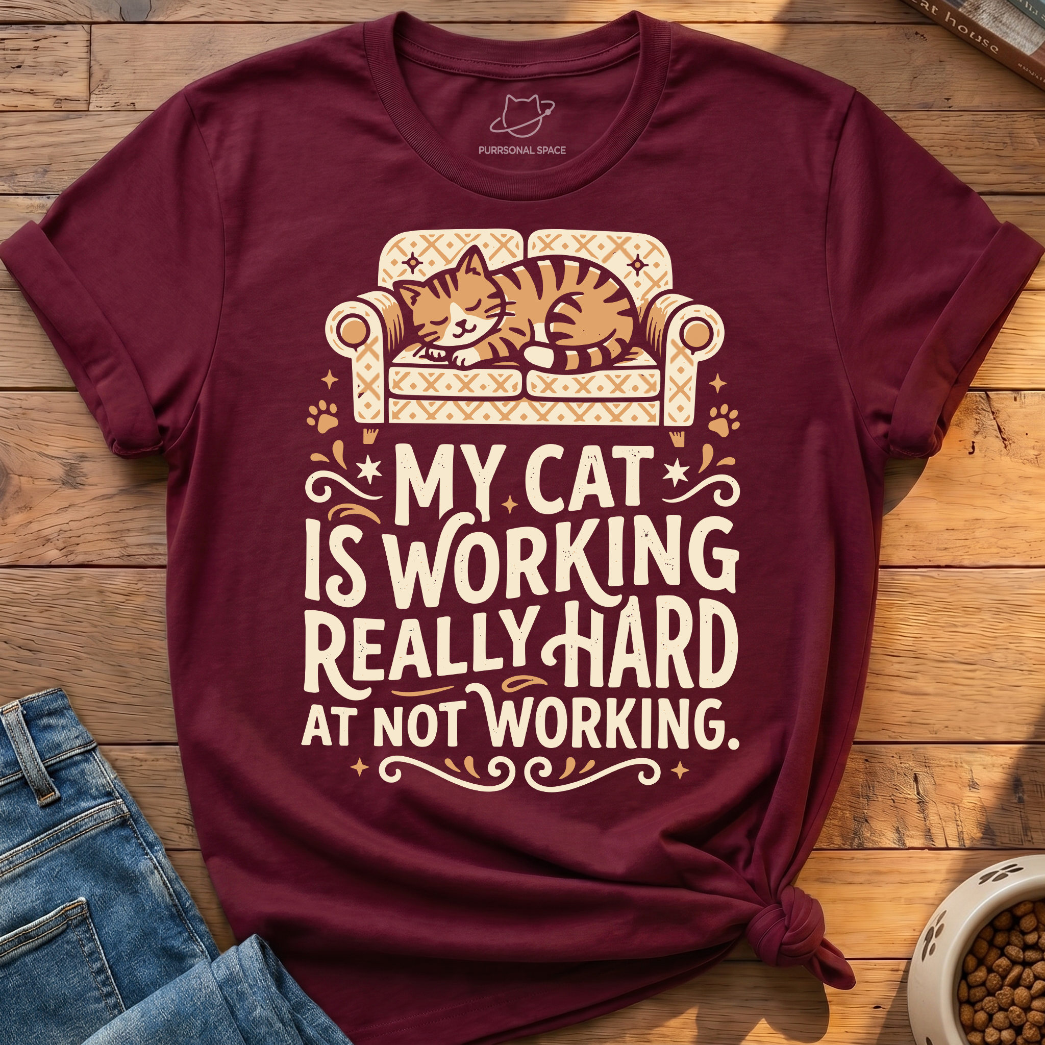 A maroon short-sleeve graphic T-shirt laid flat on a wooden surface, styled with blue jeans and a bowl of cat food. The shirt features a cream and orange illustration of a cat sleeping on a patterned sofa above the text, 'MY CAT IS WORKING REALLY HARD AT NOT WORKING.