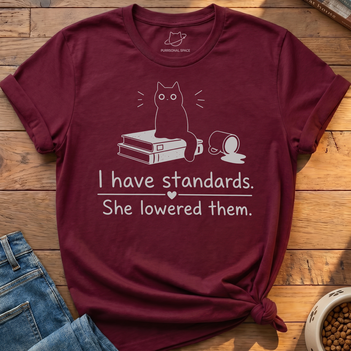 A maroon short-sleeve graphic T-shirt laid flat on a wooden surface, styled with folded blue jeans, a book, and a bowl of pet food. The shirt has a white line drawing of a cat sitting on books with a mug and the text, 'I have standards.